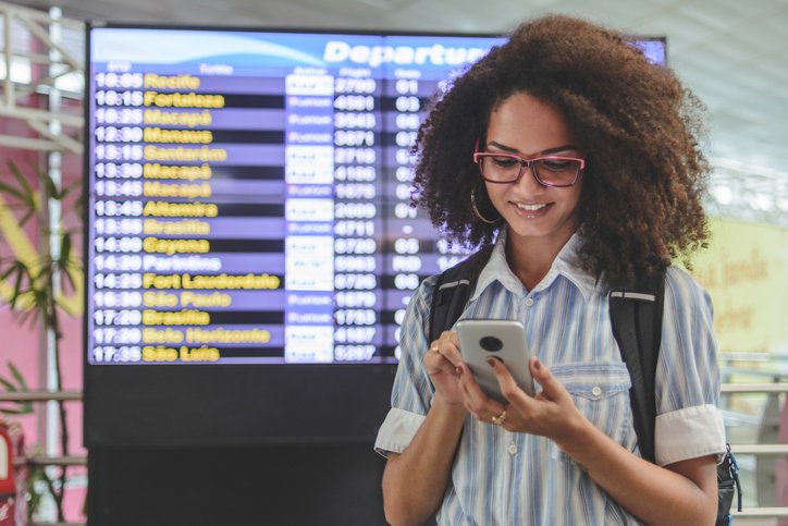 Young woman checking her boarding schedule at smartphone with airport departure time board on background